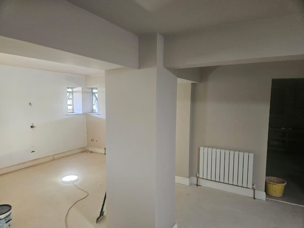 Open-plan interior painting in progress with beige walls, ceiling beams, a central pillar and a white radiator.