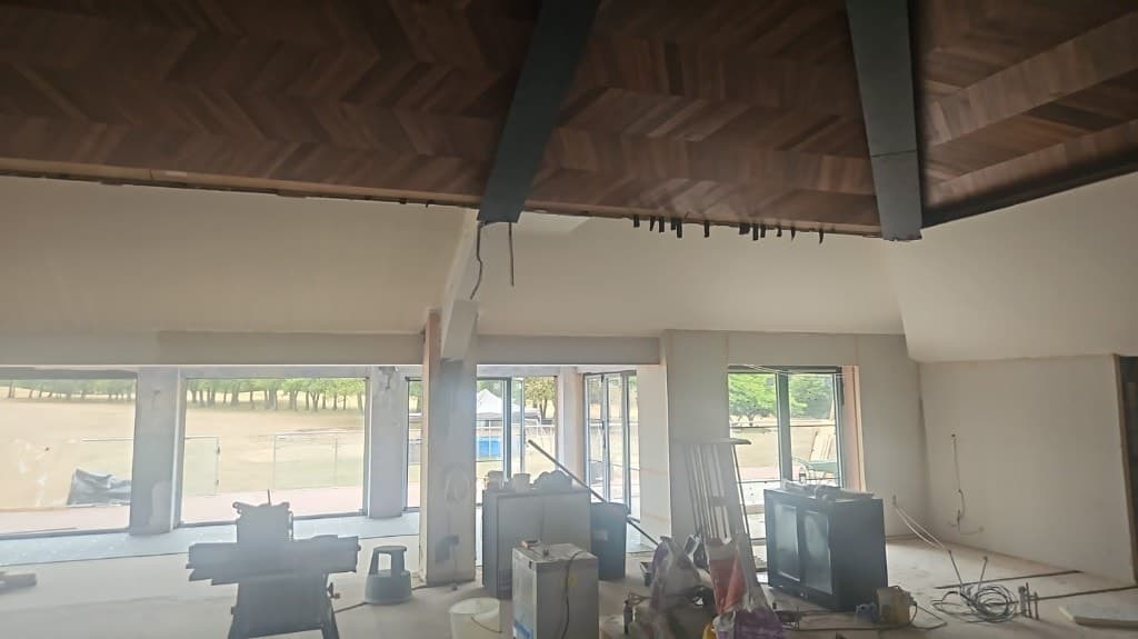 Large interior renovation with a dark wood herringbone vaulted ceiling, black structural beams, primed walls and floor-to-ceiling windows.